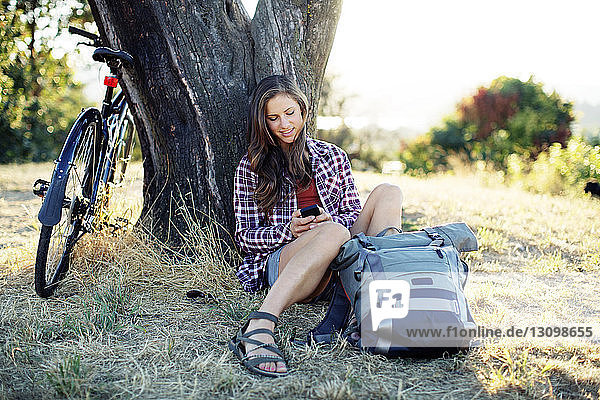Junge Frau benutzt Mobiltelefon  während sie mit Rucksack und Fahrrad auf einer Wiese sitzt
