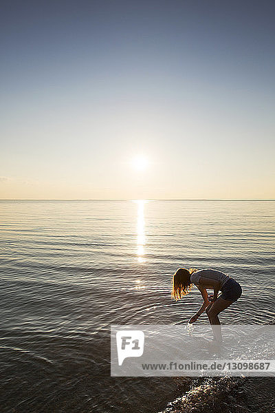 Side view of teenage girl reaching into water on shore at beach during sunset