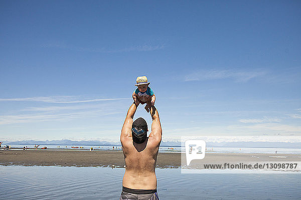 Rear view of man playing with son while standing at beach against blue sky