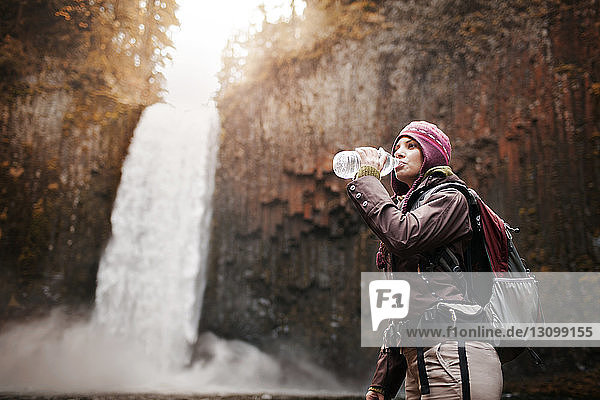 Weibliche Wanderin trinkt Wasser  während sie am Wasserfall steht