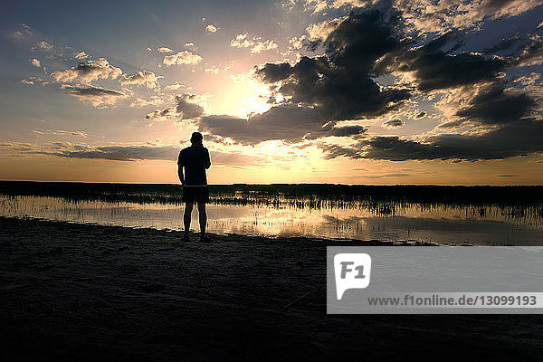 Silhouette man standing at lakeshore against cloudy sky during sunset