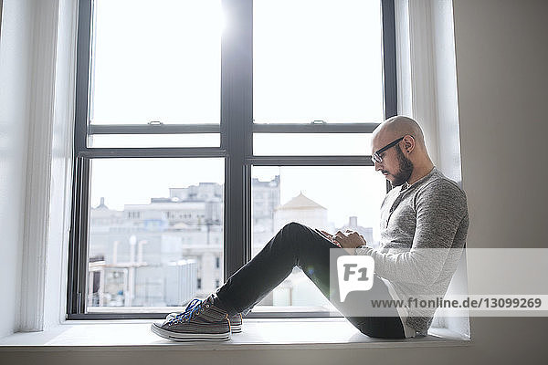 Side view of man using smart phone while sitting on window