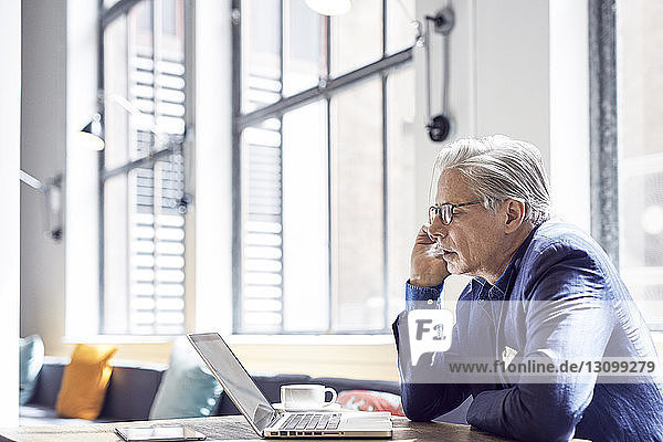 Geschäftsmann schaut auf Laptop-Computer  während er in Büro-Cafeteria an Fenstern sitzt