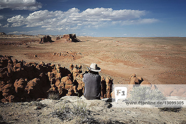 Rear view of man looking at view while siting on rock formation against sky