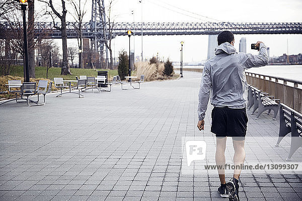 Rückansicht eines männlichen Sportlers  der die Williamsburg-Brücke beim Gehen auf einem Fußweg fotografiert