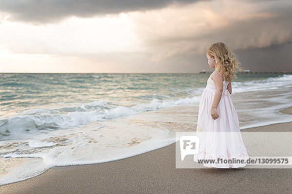 Side view of girl in dress standing at beach against cloudy sky during sunset