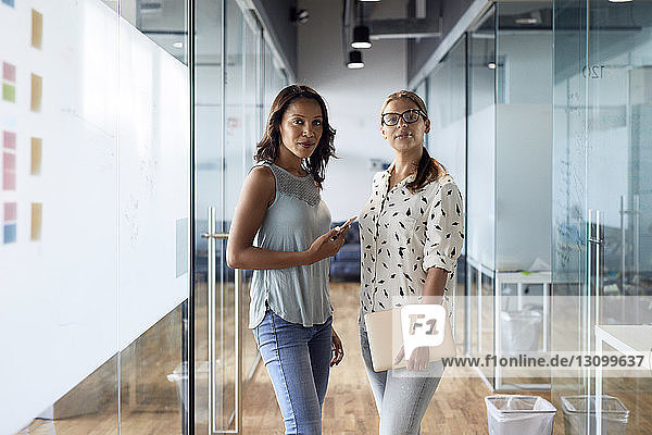 Portrait of confident businesswomen standing amidst glass walls at office