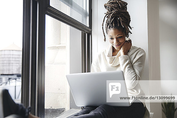 Smiling businesswoman using laptop while sitting at window sill in office