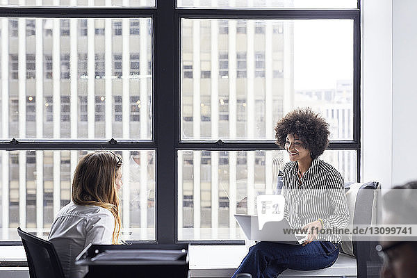 Happy businesswoman discussing with female colleague while sitting on window sill in office