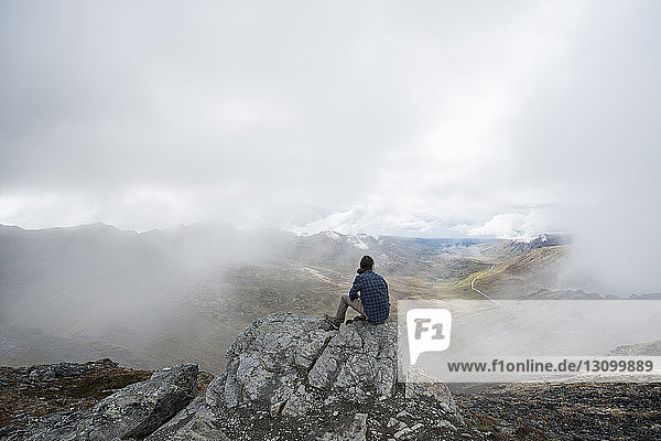 Rear view of man sitting on cliff against landscape during foggy weather