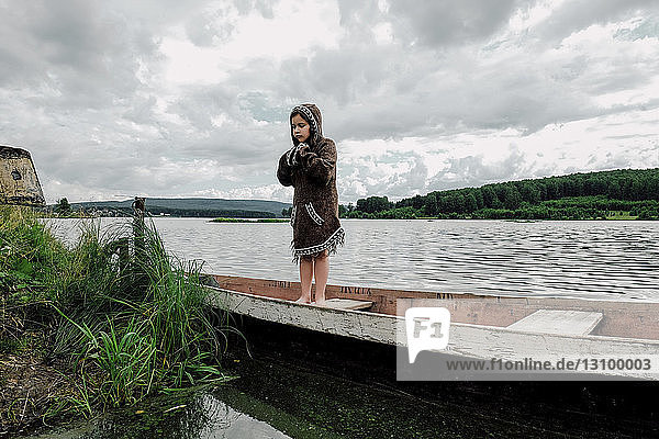 Girl wearing hooded jacket while standing in rowboat on lake