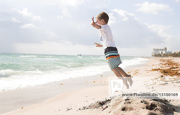 Side view of boy jumping at beach against cloudy sky