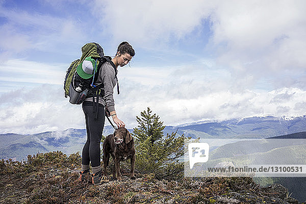 Frau steht mit Hund auf Bergfelsen vor bewölktem Himmel