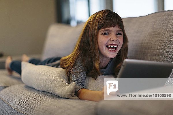 Fröhliches Mädchen benutzt Tablet-Computer  während sie zu Hause auf dem Sofa liegt