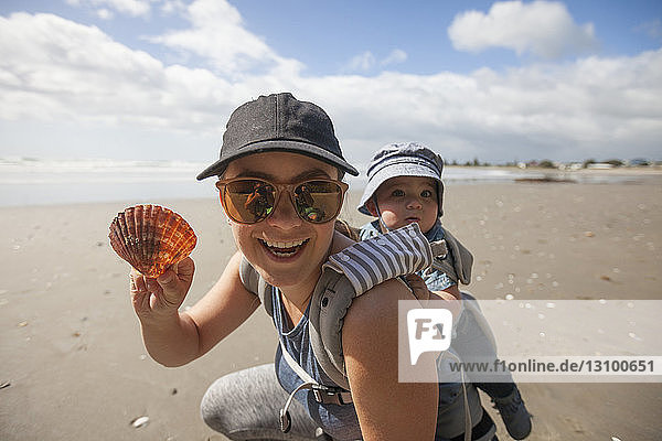 Portrait of mother holding seashell while piggybacking son at beach