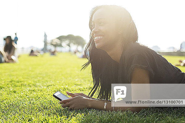 Glückliche Frau hält Mobiltelefon in der Hand  während sie bei Sonnenschein auf dem Feld im Park liegt