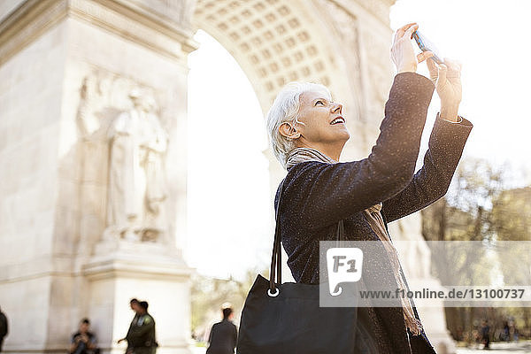 Seitenansicht einer älteren Frau  die mit einem Smartphone fotografiert  während sie in der Stadt gegen den Triumphbogen steht