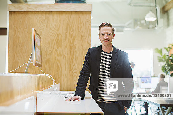 Portrait of confident businessman standing at desk by desktop computer in office