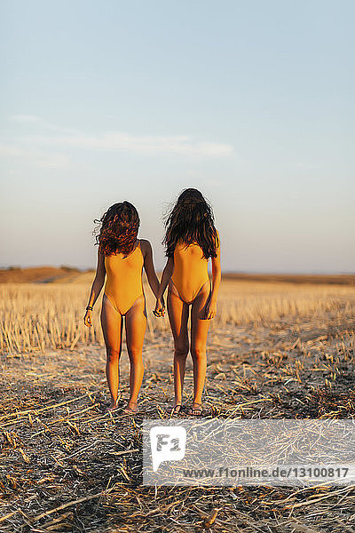 Freundinnen in Badebekleidung mit haarbedecktem Gesicht  die Hände haltend  während sie auf dem Feld gegen den Himmel stehen