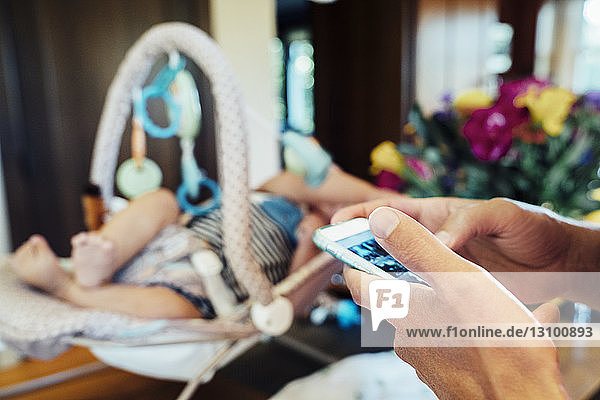 Ausgeschnittenes Bild einer Hand mit einem Smartphone von einem Baby im Kindersitz zu Hause
