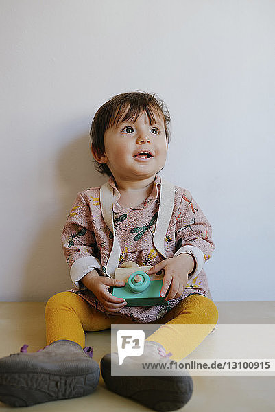 Lächelndes kleines Mädchen mit Kamera-Spielzeug  das aufschaut  während es zu Hause am Boden an der Wand sitzt
