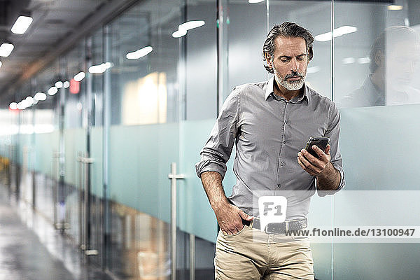Businessman with hand in pocket looking at smart phone while standing in office lobby