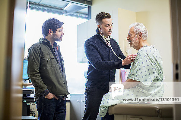 Männliche Ärzte untersuchen Patient  der auf der Krankenhausstation auf dem Bett sitzt und durch die Tür gesehen wird