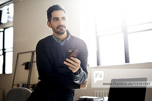 Thoughtful businessman holding smart phone and sitting in office
