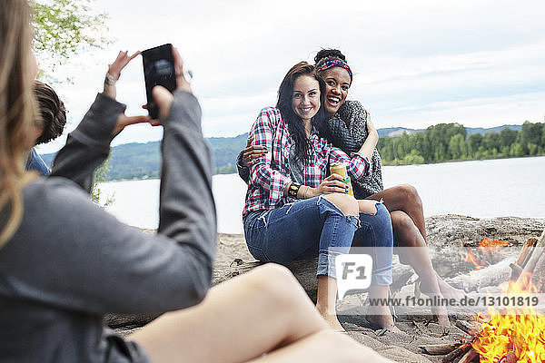 Frau fotografiert glückliche Freundinnen  die am Lagerfeuer am Fluss auf einem Baumstamm sitzen