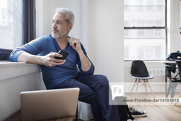 Thoughtful mature man holding smart phone while looking through window at home
