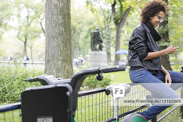 Seitenansicht einer Frau  die ein Mobiltelefon benutzt  während sie im Park auf einem Geländer sitzt