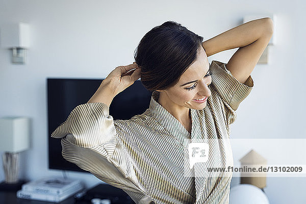 Lächelnde Frau bindet sich im Stehen zu Hause die Haare