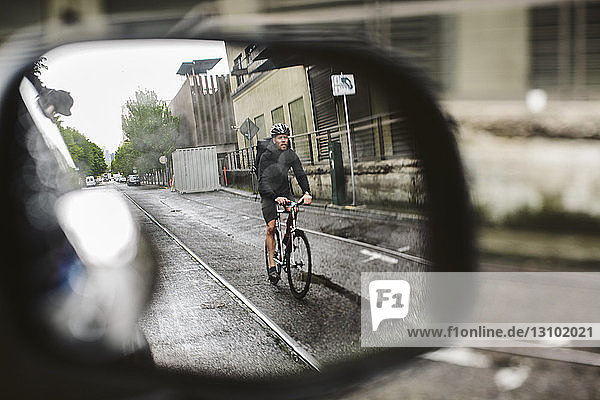 Männlicher Pendler mit Fahrrad auf nasser Straße durch den Seitenspiegel eines Autos gesehen