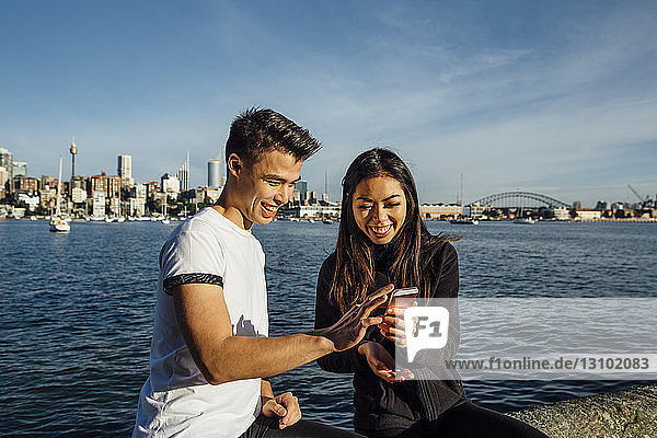Happy young couple using smart phone while exercising on promenade against Sydney Harbour Bridge