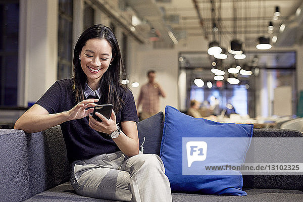 Smiling businesswoman using mobile phone while sitting on sofa at office
