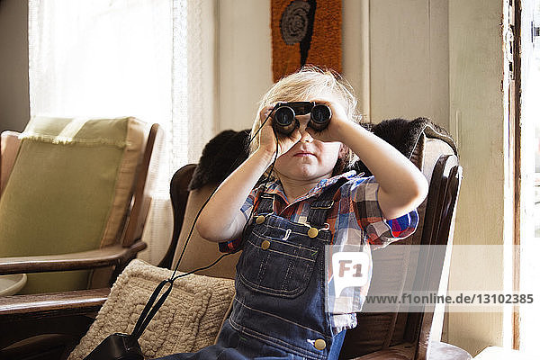 Boy looking through binoculars while sitting on chair at home