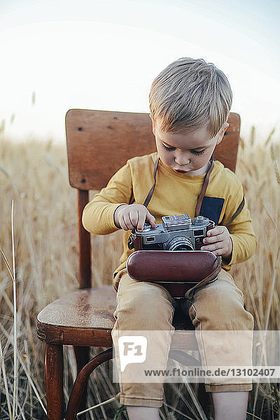 Curious boy adjusting vintage camera while sitting on chair amidst wheat field
