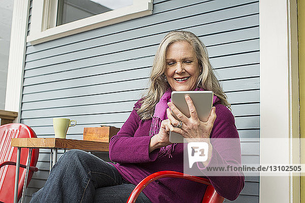 Lächelnde ältere Frau benutzt Tablet-Computer  während sie auf einem Stuhl auf der Veranda sitzt