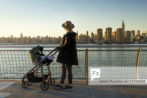 Seitenansicht einer Frau  die die Skyline der Stadt betrachtet  während sie bei Sonnenuntergang mit einem Kinderwagen auf der Promenade steht