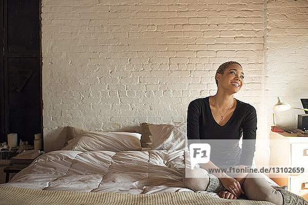 Glückliche junge Frau schaut weg  während sie zu Hause auf dem Bett sitzt