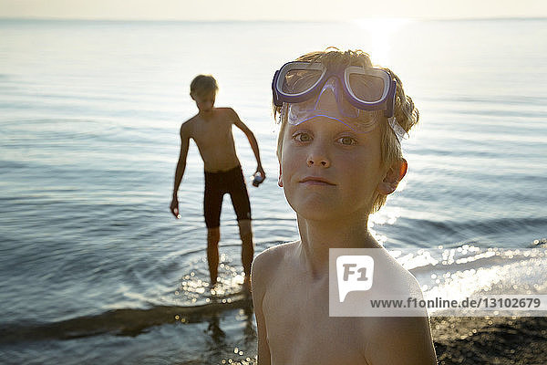 Portrait of boy wearing swimming goggles on shore with friend in background