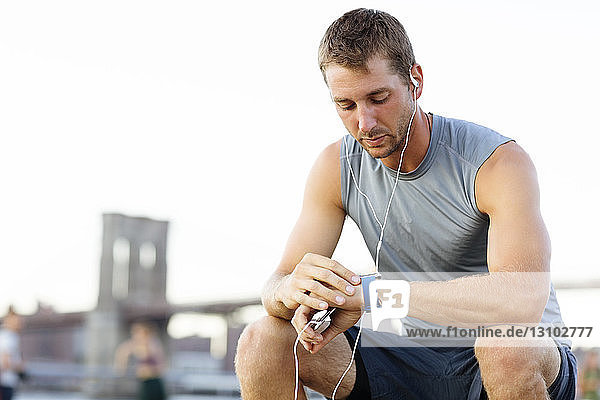 Athlete checking the time while sitting on bench against clear sky