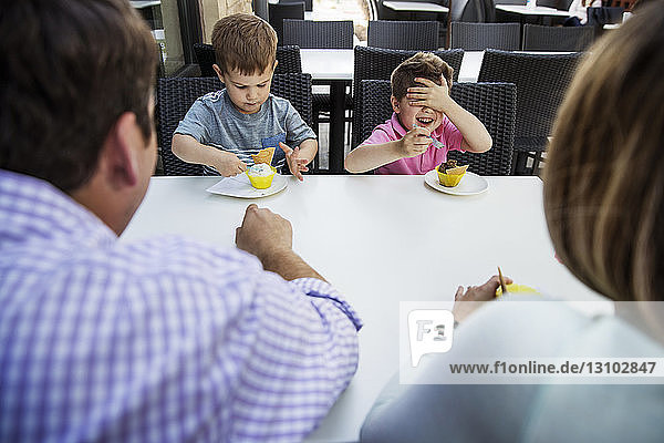 Glücklicher Junge bedeckt die Augen  während er mit seiner Familie im Restaurant Dessert isst