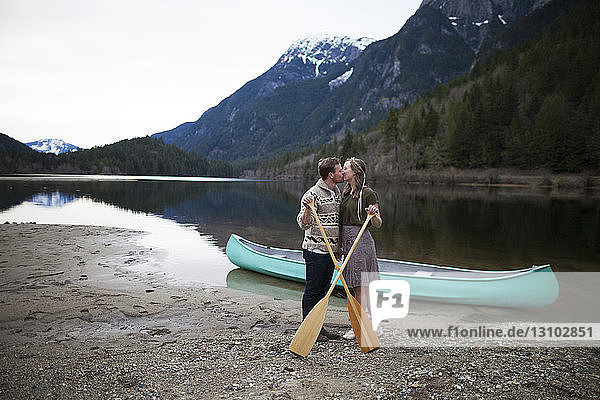 Young couple kissing while holding oars at lakeshore against mountains