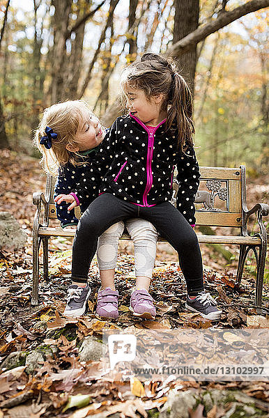 Sisters looking at each other while sitting on bench in forest during autumn