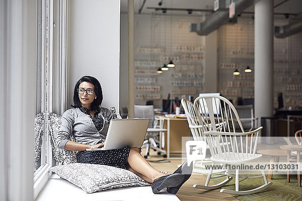 Nachdenkliche Geschäftsfrau schaut durch Fenster  während sie im Büro einen Laptop benutzt