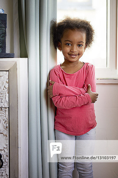 Porträt eines lächelnden Mädchens mit verschränkten Armen  das zu Hause vor einem Vorhang steht