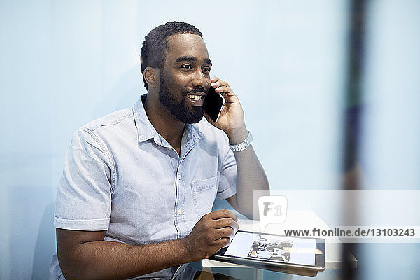 Lächelnder Geschäftsmann telefoniert mit einem Mobiltelefon  während er ein digitales Tablet in der Bürokabine benutzt