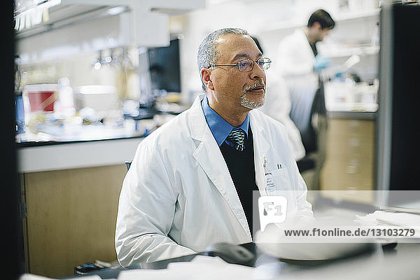 Arzt arbeitet am Schreibtisch  während ein männlicher Kollege im Hintergrund im Krankenhaus arbeitet