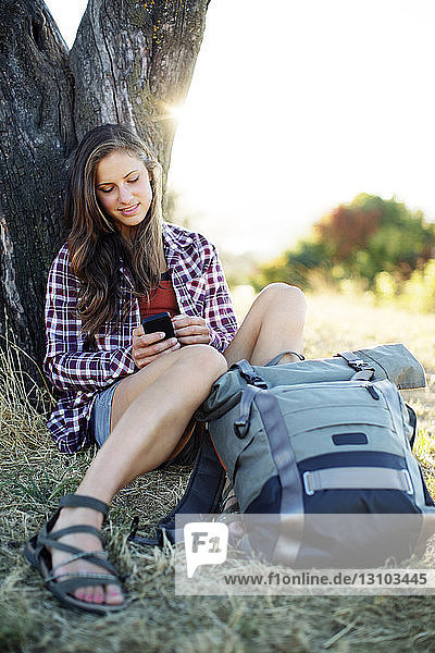 Junge Frau benutzt Mobiltelefon  während sie mit Rucksack auf einem Grasfeld sitzt